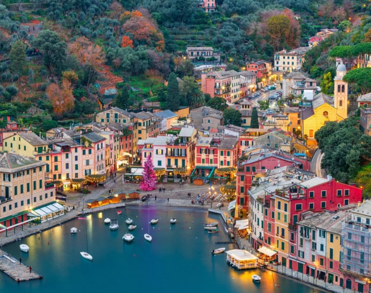Luftaufnahme des Hafens von Portofino in der Abenddämmerung mit bunten Pastellhäusern, beleuchtetem Weihnachtsbaum auf der Piazzetta und ankernden Booten.