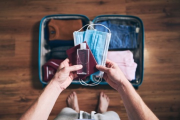 Mann hält Reisepass, Desinfektionsflasche und Corona-Schutzmasken in seinen Händen, vor im auf dem Boden ein gepackter Urlaubskoffer