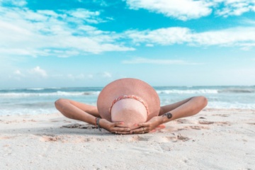 Frau mit Sonnenhut liegt am Meer im Sand und chillt