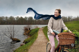 Eine Frau Fährt auf dem Fahrrad einen Weg am Ufer eines Gewässers entlang. Ihr Schal weht im Wind.