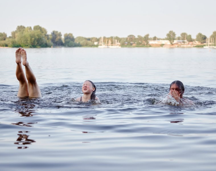 Drei Personen schwimmen in einem See, von einer sind nur die Füße und Beine zu sehen, die gerade aus dem Wasser herausgestreckt sind.