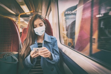 Junge Frau mit Mund-Nasen-Schutz sitzt im Zug und schaut auf Handy