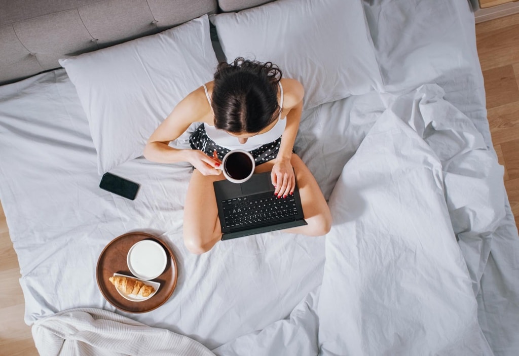 Effiziente junge Millennial Mädchen sitzt auf einem Bett am Morgen, verwendet Laptop-Computer und isst Croissants und trinkt Kaffee zum Frühstück. Top Down Shot.