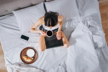 Effiziente junge Millennial Mädchen sitzt auf einem Bett am Morgen, verwendet Laptop-Computer und isst Croissants und trinkt Kaffee zum Frühstück. Top Down Shot.