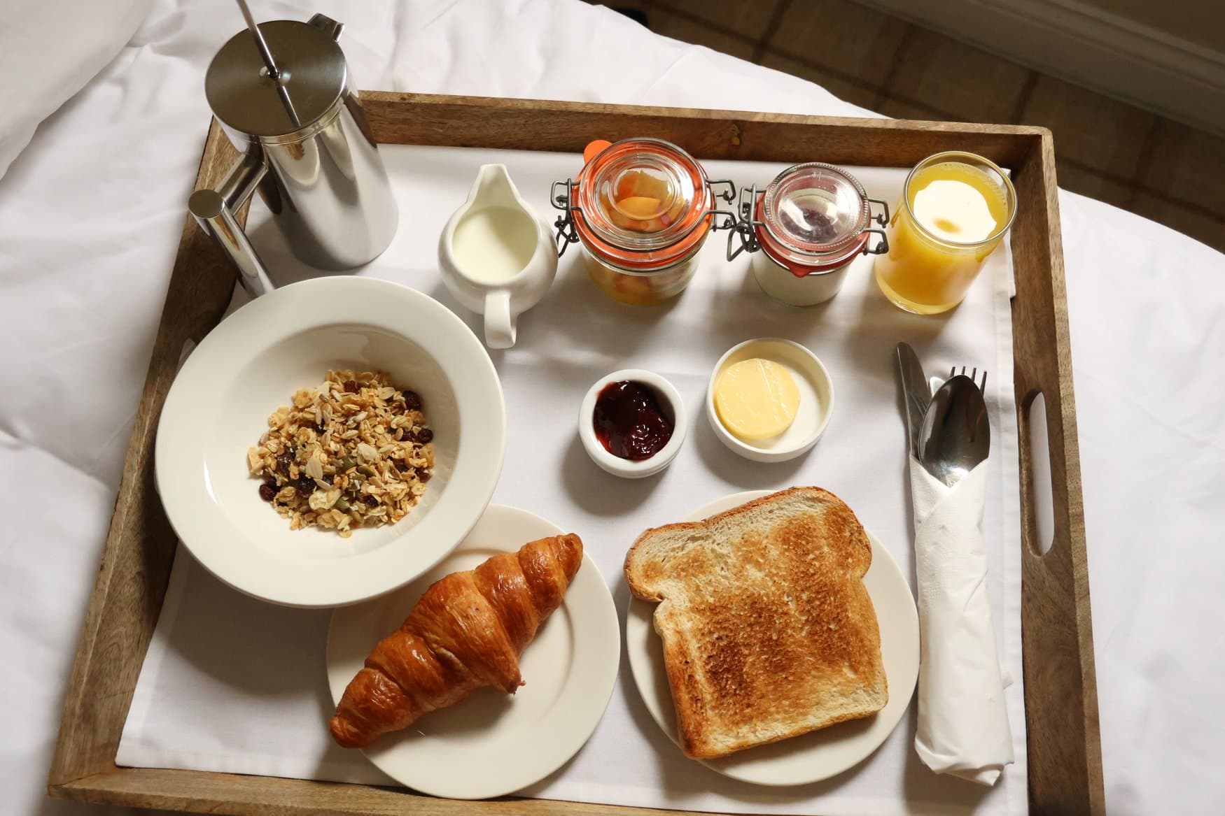 Blick von oben auf ein Frühstückstablett aus Holz, das auf einem Bett steht. Darauf Kaffeekanne, Müsli, Croissant, Toast, Marmelade und Orangensaft.