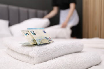 Zwei Euro-Scheine stecken zwischen frischen Handtüchern auf einem Hotelbett, im unscharfen Hintergrund richtet eine Zimmermitarbeiterin die Kissen.