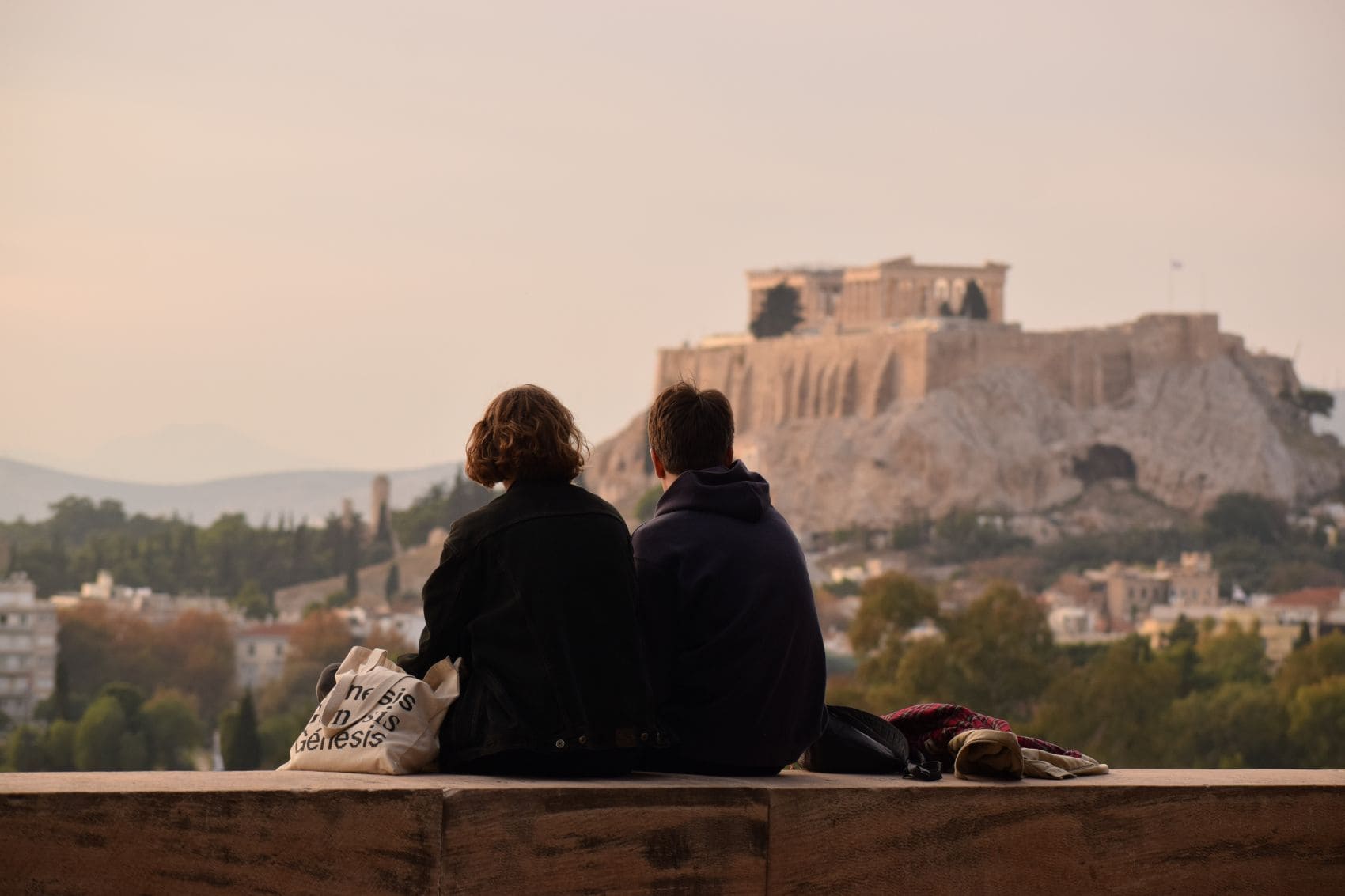 Junger Mann und junge Frau sitzen auf Mauer und genießen Aussicht auf Athen 