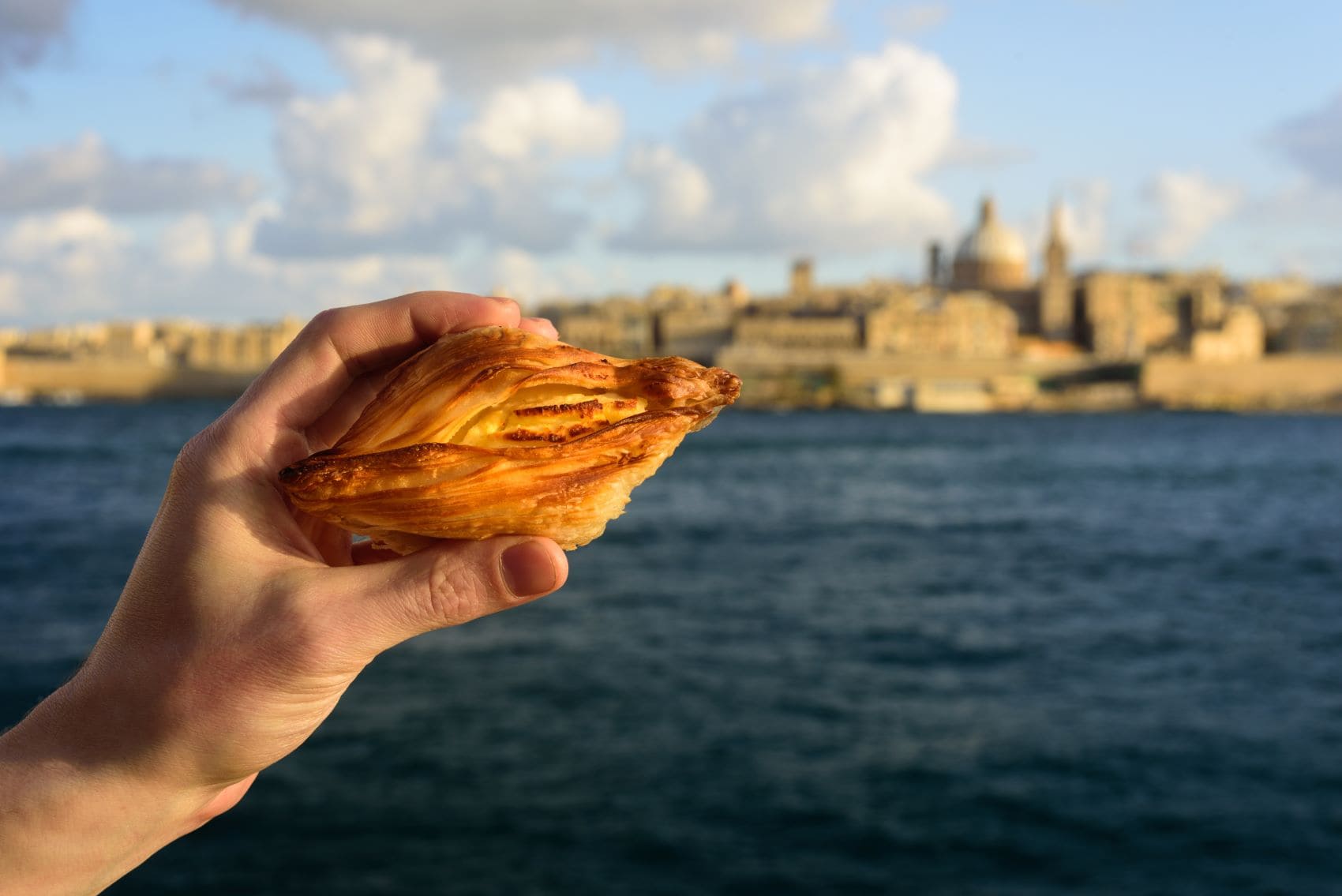 Person hält Pastizzi in der Hand, im Hintergrund Panorama von Valletta 