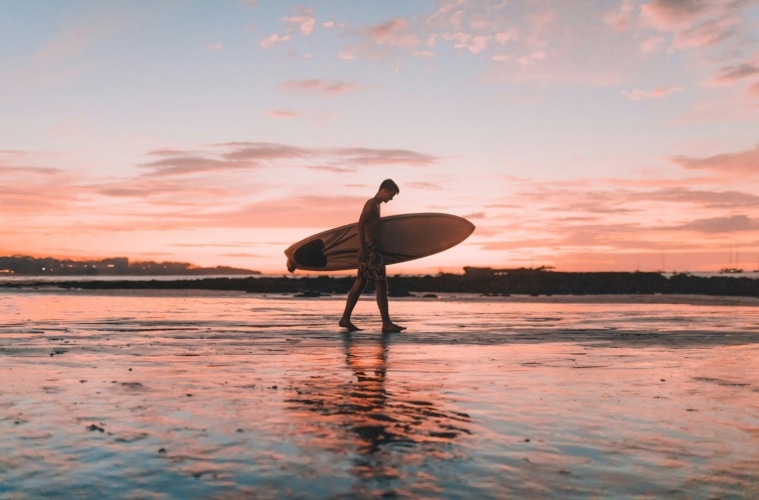Mann mit Surfbrett bei Sonnenuntergang am Strand