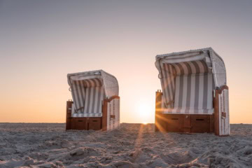 Zwei Strandkörbe an einem Strand an der Nordsee in Nahaufnahme, dahinter geht die Sonne im Meer unter.