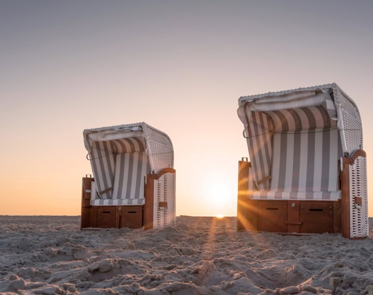 Zwei Strandkörbe an einem Strand an der Nordsee in Nahaufnahme, dahinter geht die Sonne im Meer unter.