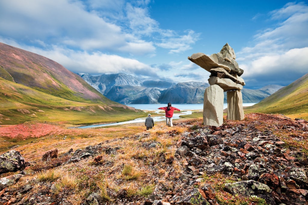 Ein Steindenkmal der Inuit steht im Torngat Nationalpark, dahinter läuft eine ältere Person und ein Kind, das das Steindenkmal imitiert.