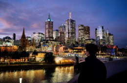 Eine erleuchtete Skyline vor einem nachtblauen Himmel. Im Vordergrund steht eine Person mit dem Rücken zum Betrachter auf einer Brücke und blickt auf die Skyline. Der Fluss zwischen den Ufern ist von den Lichtern der Stadt erleuchtet.