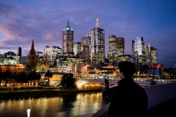 Eine erleuchtete Skyline vor einem nachtblauen Himmel. Im Vordergrund steht eine Person mit dem Rücken zum Betrachter auf einer Brücke und blickt auf die Skyline. Der Fluss zwischen den Ufern ist von den Lichtern der Stadt erleuchtet.