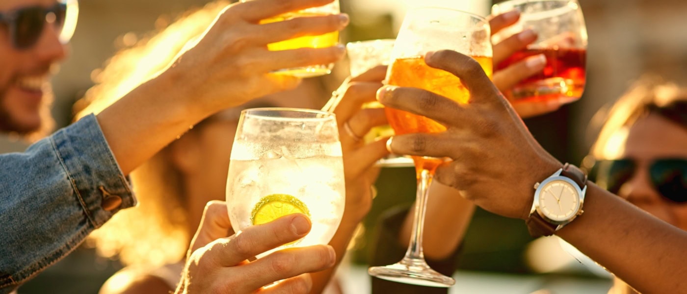 Mehrere Menschen stoßen mit sommerlichen Drinks in der Sonne miteinander an.
