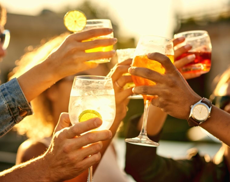 Mehrere Menschen stoßen mit sommerlichen Drinks in der Sonne miteinander an.