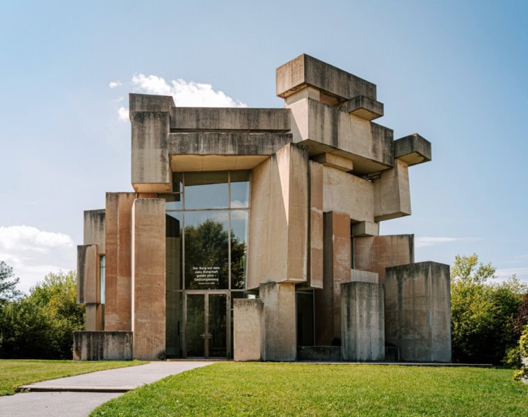 Auf einem grünen Hügel steht eine brutalistische Kirche, die aus zahlreichen großen Betonquadern besteht. Das Einlassportal ist in eine mehrere Meter hohe Glasfront eingelassen.