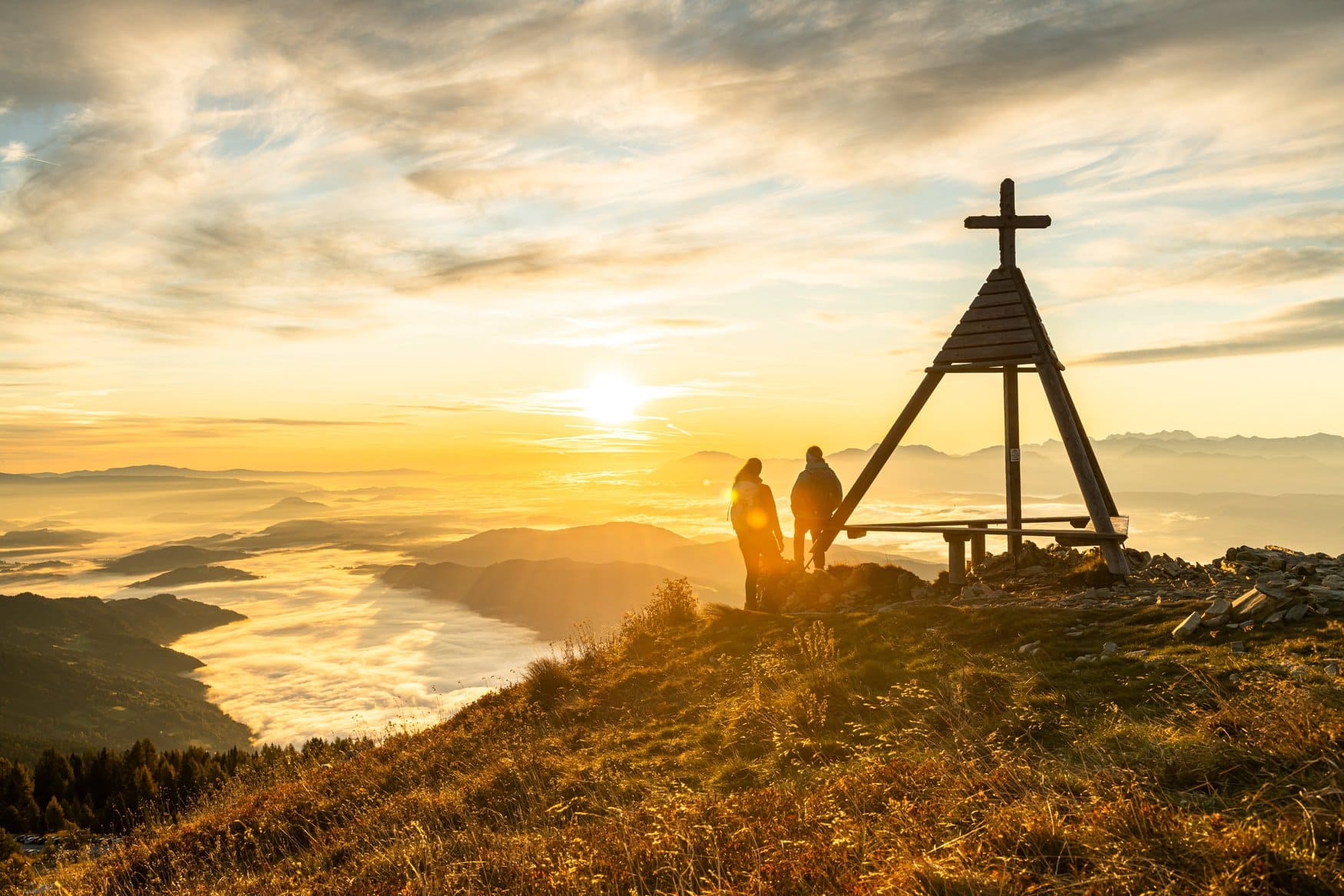 Zwei Wandernde stehen bei Sonnenaufgang neben dem Gipfelkreuz der Gerlitzen. Unter ihnen liegt ein Nebelmeer, aus dem die Bergrücken herausragen.