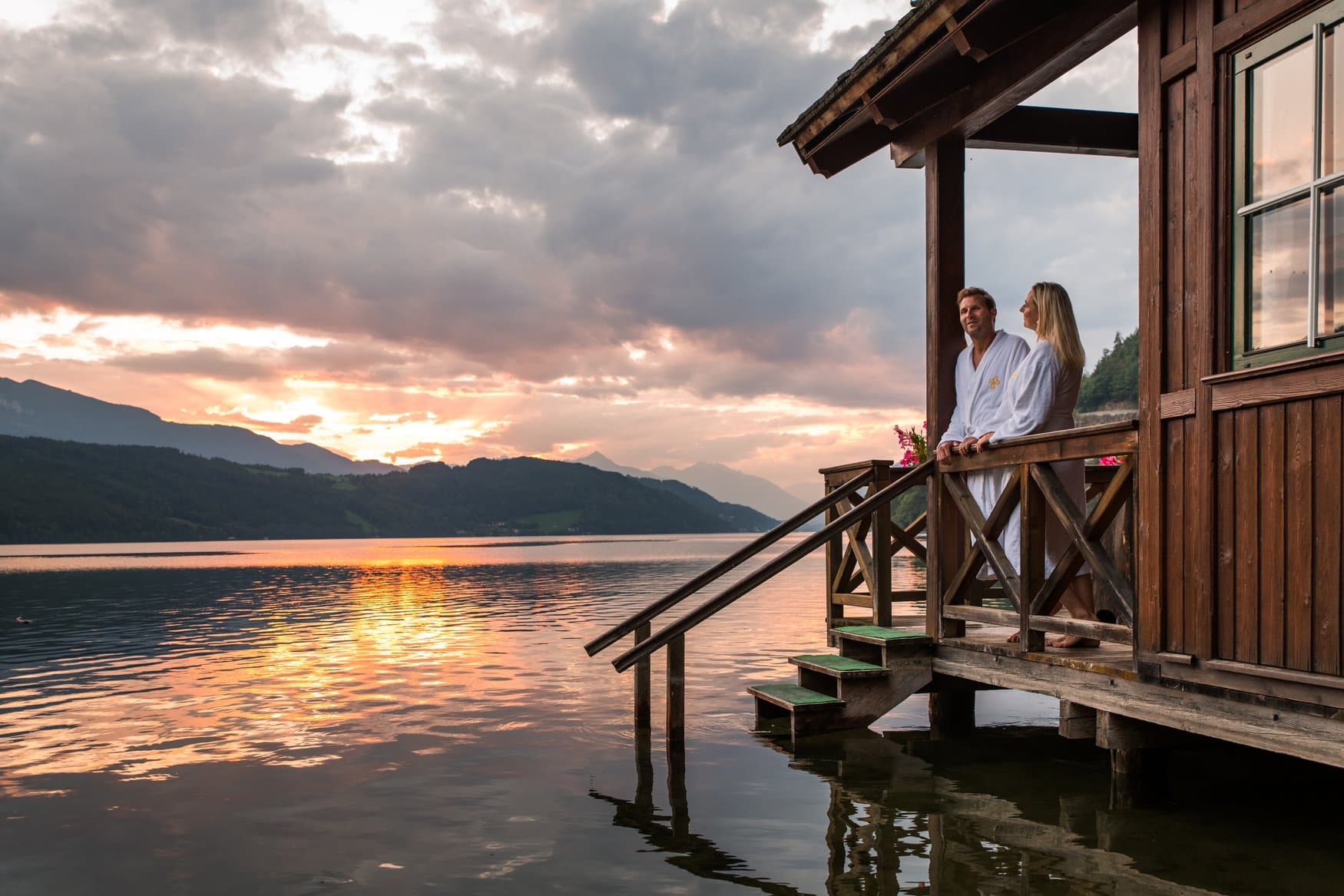 Ein Paar in weißen Bademänteln steht auf der Terrasse der Seesauna Seefischer am Millstätter See und blickt auf den Sonnenuntergang über den Bergen.