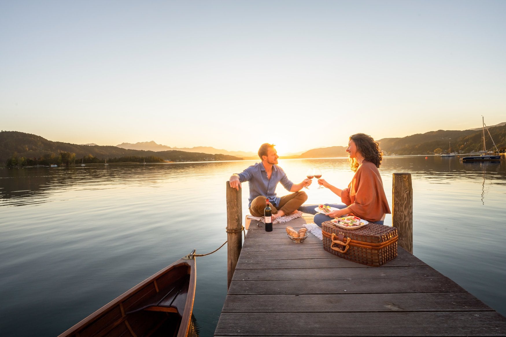 Ein Paar sitzt bei Sonnenuntergang mit einem Picknickkorb auf einem Holzsteg am Wörthersee bei Pörtschach und stößt mit Rotwein an. Links liegt ein hölzernes Ruderboot vertäut.