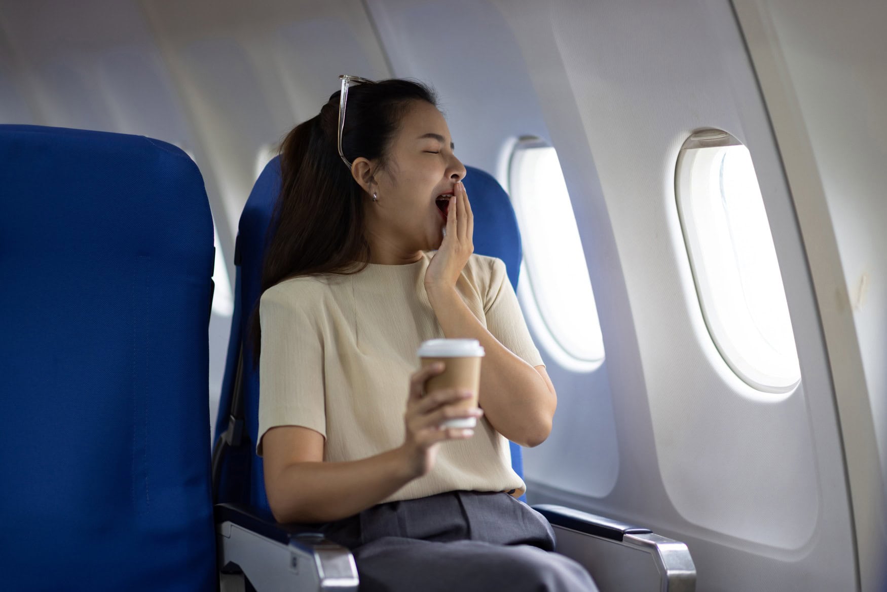 Eine junge Frau sitzt in einem Flugzeug, gähnt und hält sich dabei die Hand vor den Mund.
