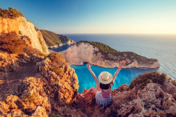 Frau mit Sonnenhut und ausgebreiteten Armen blickt auf Küste der Insel Zakynthos, Griechenland