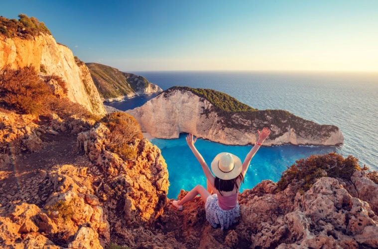 Frau mit Sonnenhut und ausgebreiteten Armen blickt auf Küste der Insel Zakynthos, Griechenland