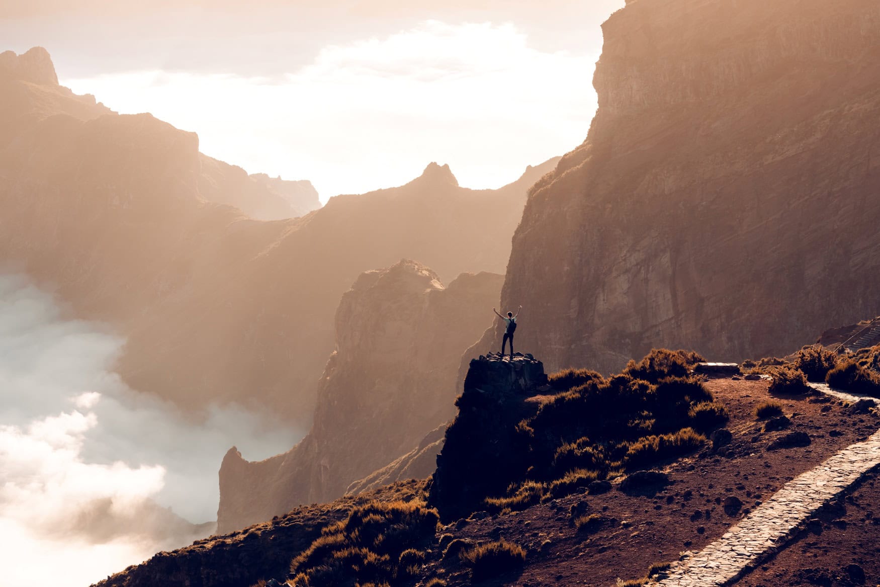 Eine Person steht am Rand eines gepflasterten Wanderwegs auf Madeira und blickt in ein zerklüftetes Tal, in dem einige Wolken hängen.
