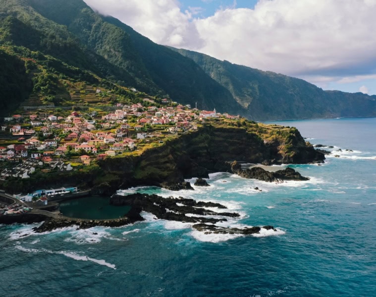 Panoramaaufnahme eines Dorfs an einer Klippe von Madeira; links die Insel mit Gebäuden, rechts der Atlantik.