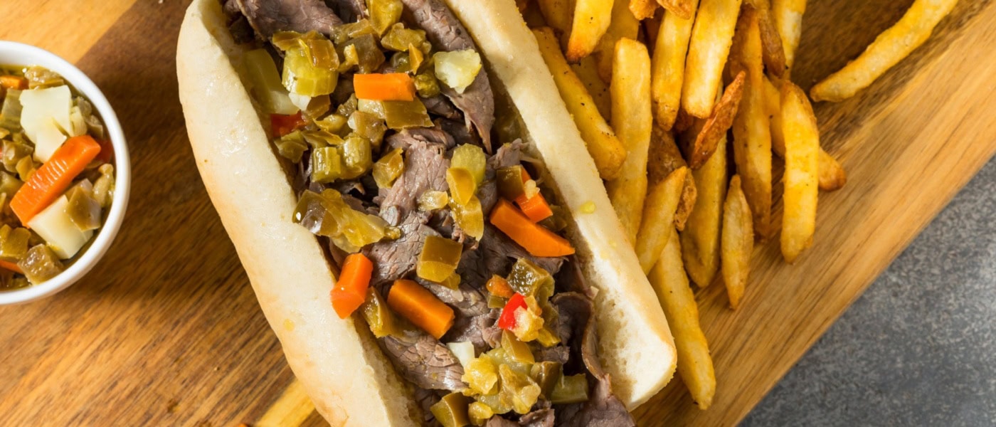 Auf einem Holzbrett liegt ein Italian Beef Sandwich Chicago Style, daneben eine Portion Pommes.