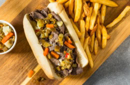 Auf einem Holzbrett liegt ein Italian Beef Sandwich Chicago Style, daneben eine Portion Pommes.