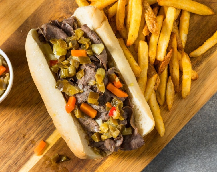 Auf einem Holzbrett liegt ein Italian Beef Sandwich Chicago Style, daneben eine Portion Pommes.