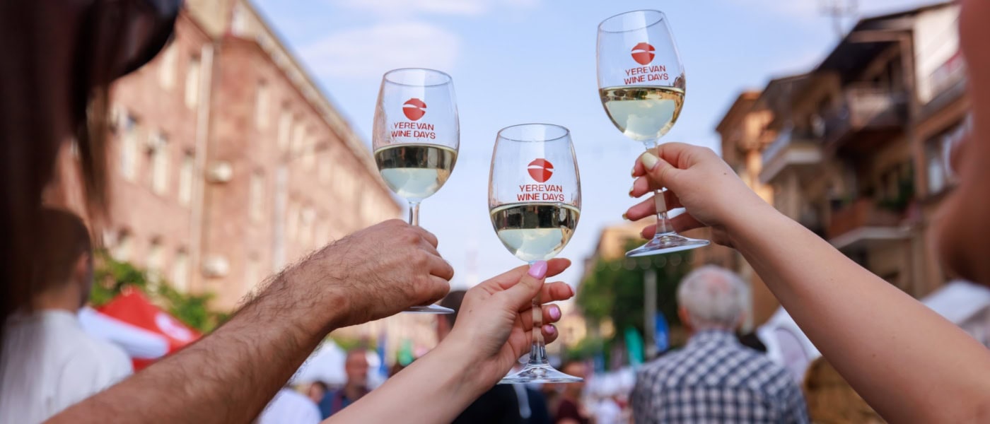 Drei Hände stoßen mit drei Weingläsern, jeweils mit der Aufschrift "Yerevan Wine Days" an; unscharf dahinter eine Szene auf einem Straßenfest in Armenien.