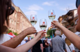Drei Hände stoßen mit drei Weingläsern, jeweils mit der Aufschrift "Yerevan Wine Days" an; unscharf dahinter eine Szene auf einem Straßenfest in Armenien.