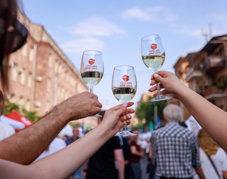 Drei Hände stoßen mit drei Weingläsern, jeweils mit der Aufschrift "Yerevan Wine Days" an; unscharf dahinter eine Szene auf einem Straßenfest in Armenien.