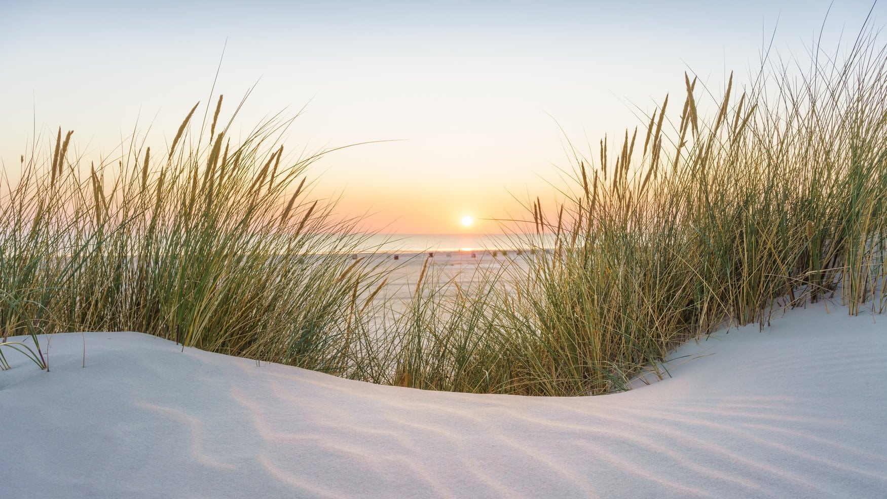 Blick über eine Sanddüne mit Strandhafer auf die Nordsee bei Sonnenuntergang, im Hintergrund stehen Strandkörbe am Wassersaum.