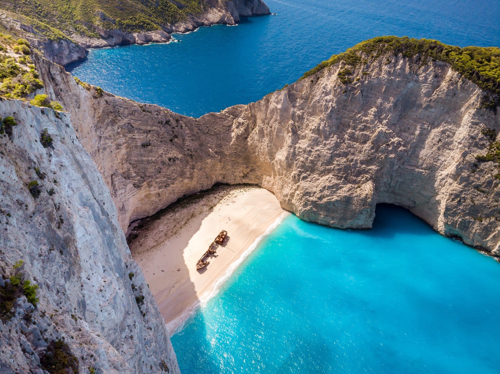 Blick von der Klippe auf die Bucht von Navagio auf Zakynthos: ein rostiges Schiffswrack liegt im hellen Sand, umgeben von steilen Kalksteinwänden und türkisfarbenem Meer.