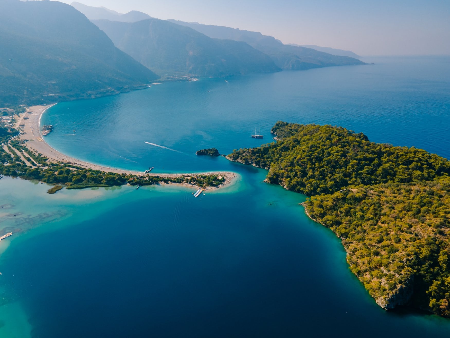 Drohnenaufnahme der Blauen Lagune von Ölüdeniz bei Fethiye mit der bewaldeten Halbinsel, der schmalen Sandzunge und dem langen Strand vor dem Küstengebirge.