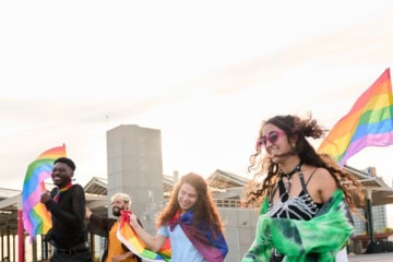 Mehrere junge Menschen mit Regenbogenflaggen rennen über einen offenen Platz und haben sichtlich gute Laune.