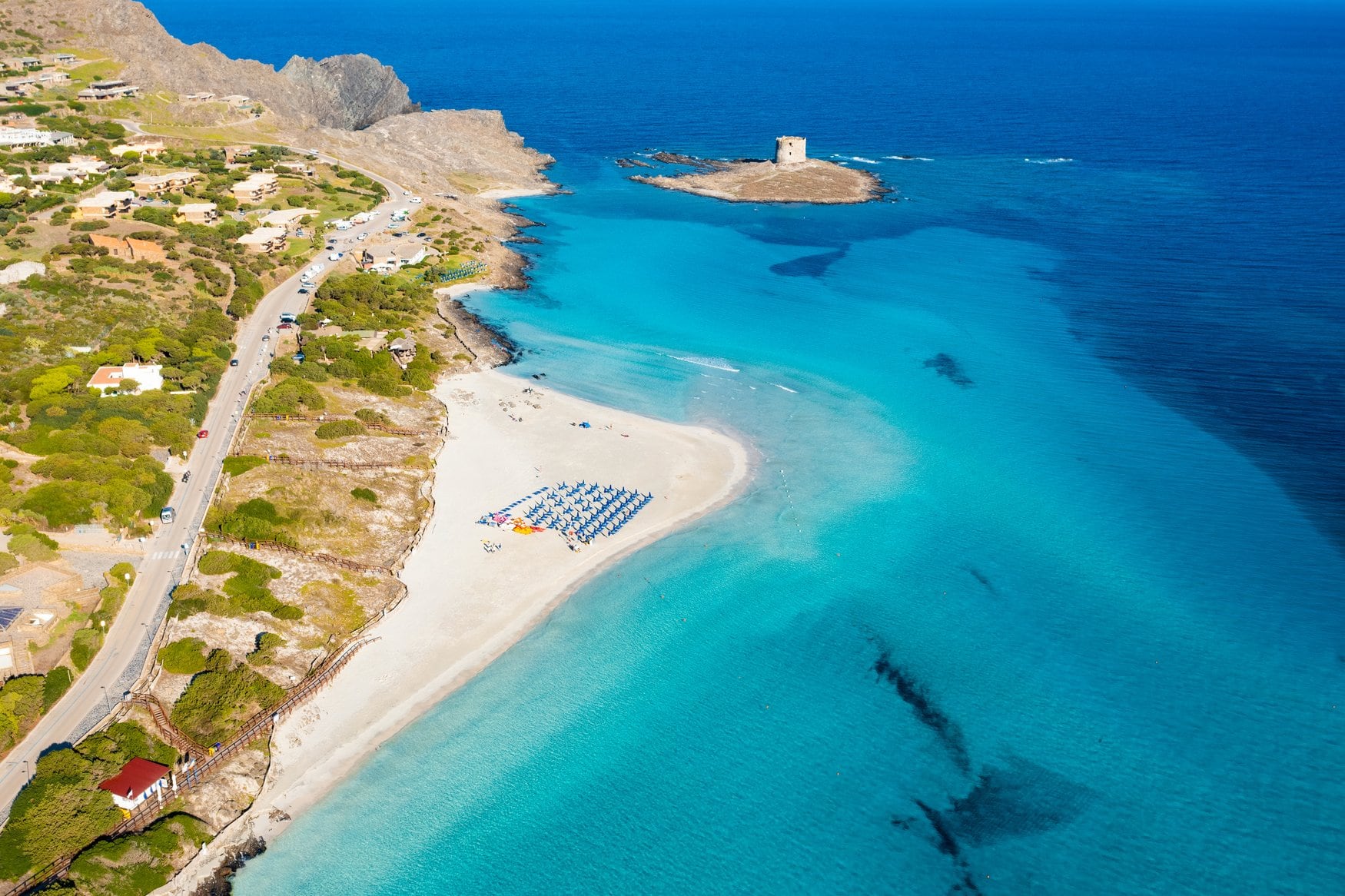 Luftaufnahme des Strands La Pelosa bei Stintino im Nordwesten Sardiniens mit flachem türkisfarbenem Wasser, Sonnenschirmreihen am hellen Sand und dem Wachturm auf der vorgelagerten Felseninsel.