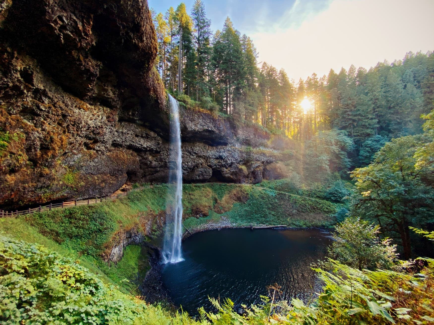 Ein schmaler Wasserfall läuft über eine rote Klippe und in einen Waldsee. Zwischen den Bäumen scheint die Sonner hervor.
