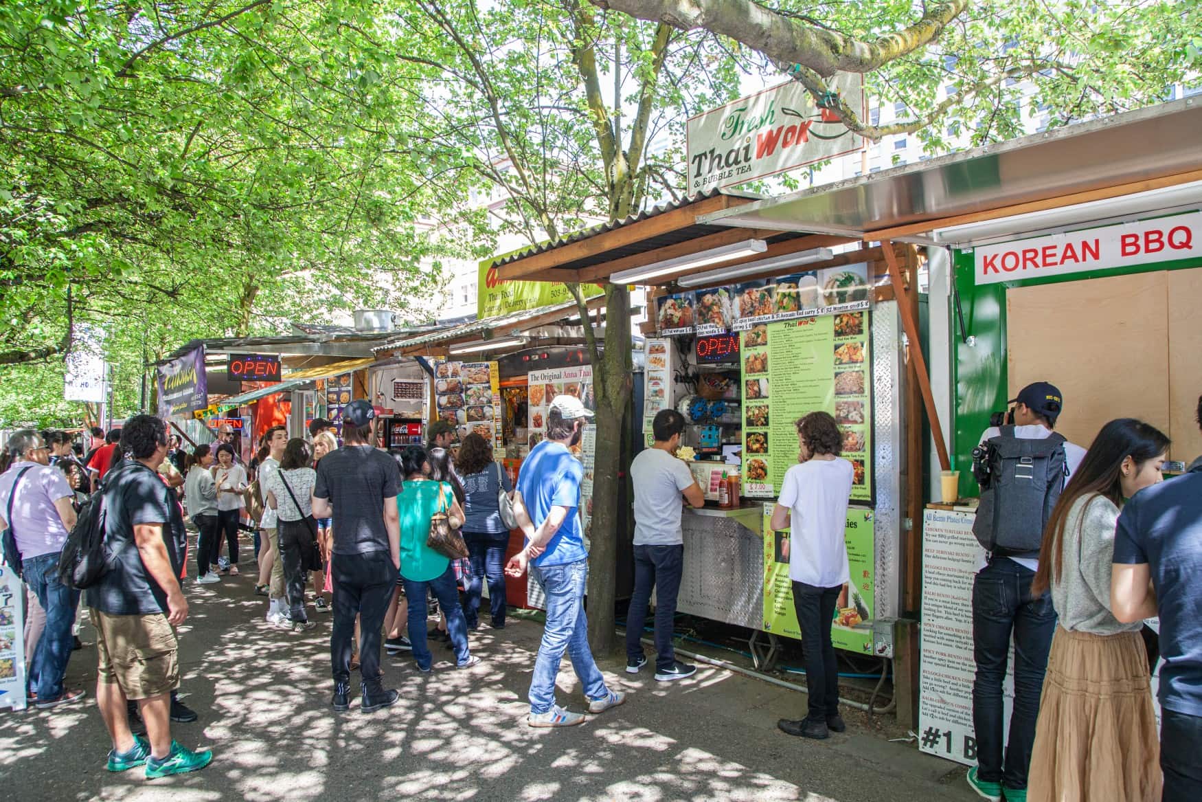 Vor einer Reihe an Food Trucks in einer sommerlichen Allee stehen zahlreiche Menschen in mehreren Schlangen an.