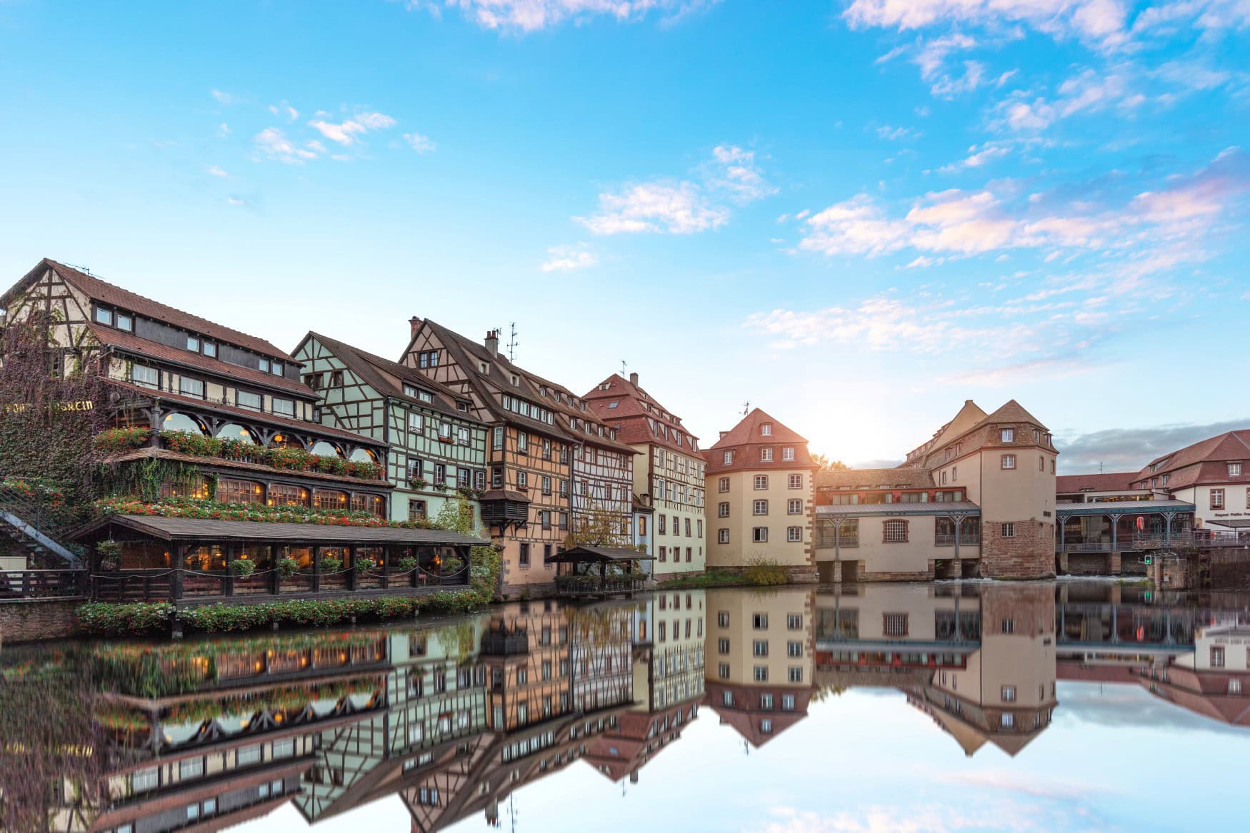Entlang eines Kanals in Straßburg reihen sich mehrere Fachwerkhäuser dicht an dicht, deren Fassaden sich im glatt liegenden Wasser perfekt spiegeln.