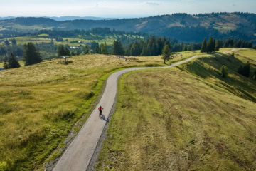 Drohnenaufnahme einer Person, die auf einem langezogen Radweg entlang einer grüner Hügel im Allgäu unterwegs ist.