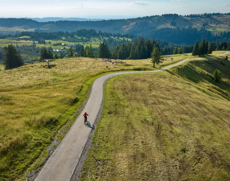 Drohnenaufnahme einer Person, die auf einem langezogen Radweg entlang einer grüner Hügel im Allgäu unterwegs ist.