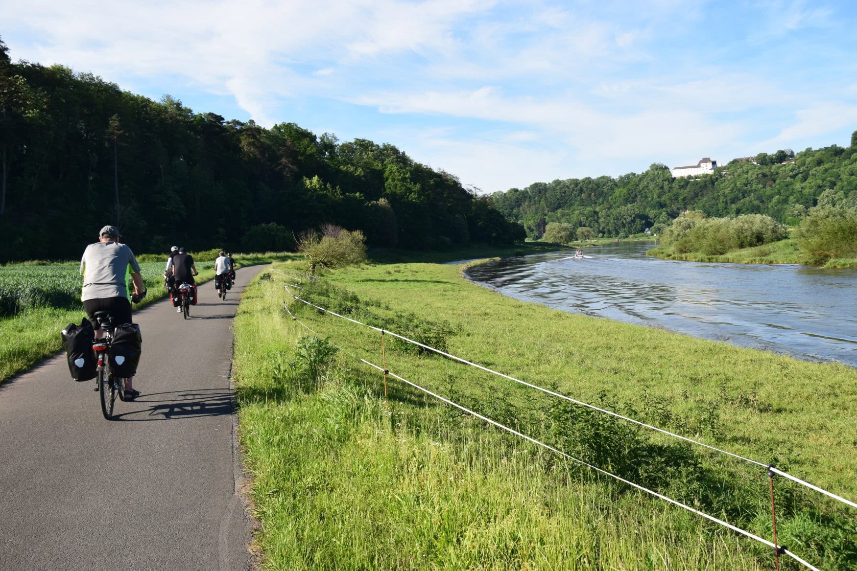 Vom Betrachter weg führt ein breiter Fluss Richtung Horizont, bevor er um eine Linksbiegung verschwindet. Links des Flusses führt ein Radweg entlang, auf dem drei Menschen entlang radeln.