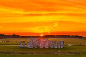 Luftaufnahme von Stonehenge in England; im Hintergrund geht über einem Waldstück rot-gelb die Sonne unter.