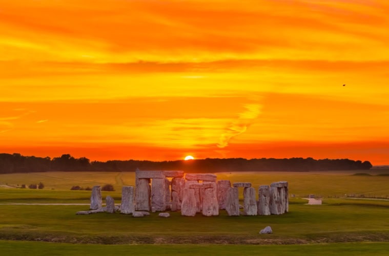 Luftaufnahme von Stonehenge in England; im Hintergrund geht über einem Waldstück rot-gelb die Sonne unter.