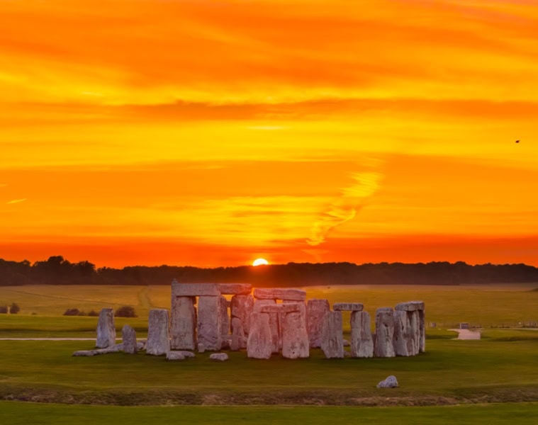 Luftaufnahme von Stonehenge in England; im Hintergrund geht über einem Waldstück rot-gelb die Sonne unter.
