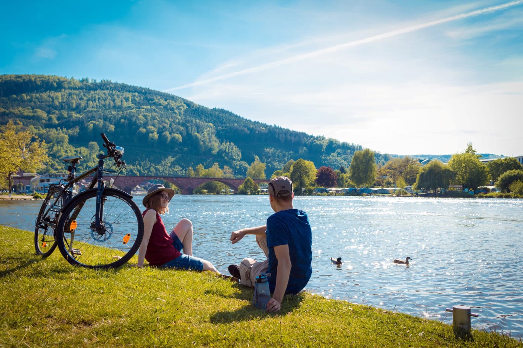 Zwei Menschen sitzen in der Sonne am Ufer des Mains, im Hintergrund eine Brücke. Neben ihnen steht ein Fahrrad.